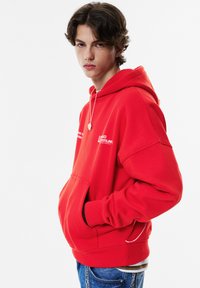 Punainen huppari, jossa on löysä istuvuus, etutasku ja valkoiset logoyksityiskohdat. Valmistettu pehmeästä kankaasta, jossa on resorit hihansuissa, yhdistettynä sinisiin farkkuihin.