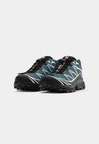 Trailrunning schoenen in teal en zwart met een gestructureerde Gore-Tex bovenkant, prominente witte accenten en een robuuste, gripvaste buitenzool.
