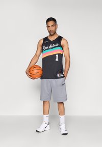 Juoda krepšinio apranga su užrašu „San Antonio“ ir daugiataučiais horizontaliais dryžiais, derinama su pilkomis trumpomis kelnėmis ir baltais sportbačiais, laiko krepšinį.