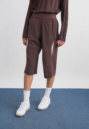 Άτομο που φορά φαρδιά καφέ βερμούδα μέχρι το γόνατο με μπεζ πλευρικές πινελιές, συνδυασμένη με λευκές κάλτσες και λευκά αθλητικά παπούτσια, στέκεται σε μπλε χαλί.