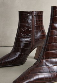 Dunkelbraune Stiefeletten aus Leder mit Krokodilprägung, seitlichem Reißverschluss und Blockabsätzen auf neutralem Hintergrund.