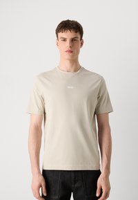 Νέος άνδρας που φοράει μπεζ T-shirt με το λογότυπο BOSS και σκούρο παντελόνι, στέκεται μπροστά σε απλό ανοιχτό γκρι φόντο.