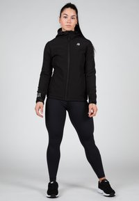 Melnā kapucēta jaka ar rāvējslēdzēju, pieguļoša piegriezuma un pelēku logo. Saskaņota ar melniem leggiem un melnām sporta apaviem. Gluda tekstūra.