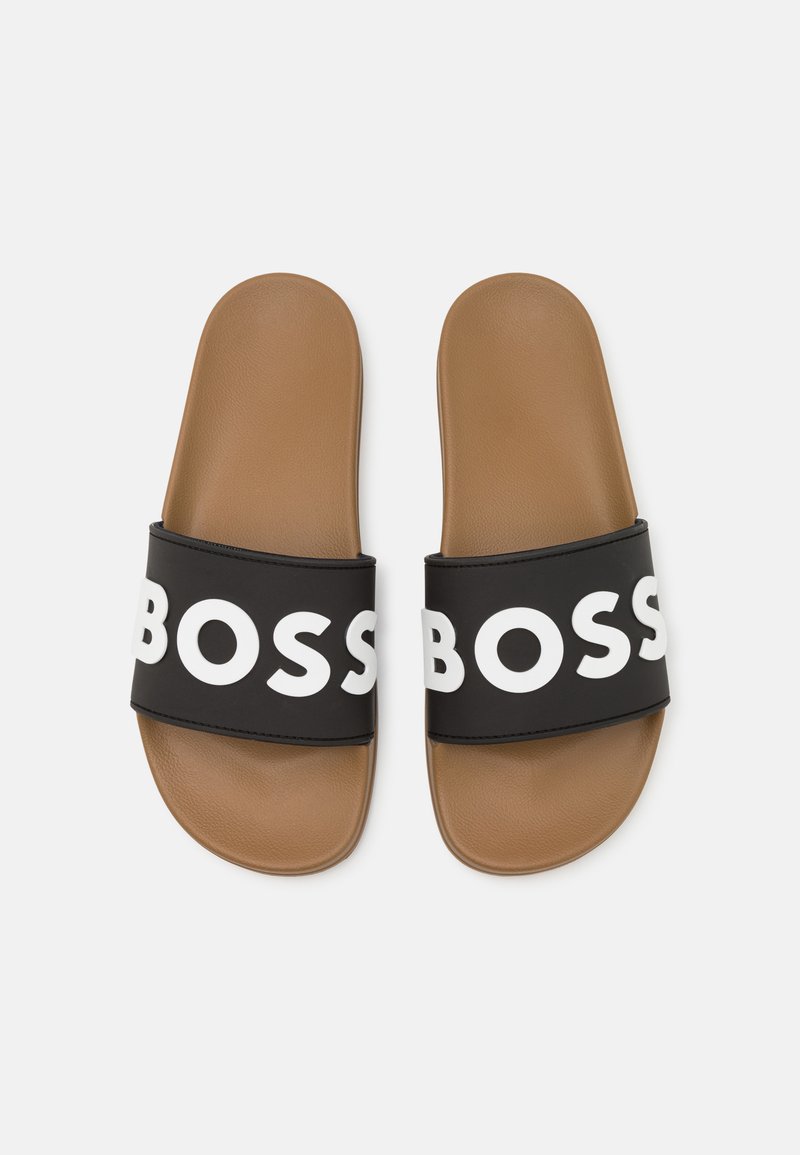 Des sandales noires à glissière avec une large lanière portant le texte blanc « BOSS ». L'intérieur est brun, fait d'un matériau lisse avec une forme arrondie.