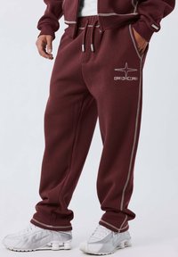Pantalones de chándal burdeos con rayas laterales y logo. Hechos de tela suave, presentan una cinturilla elástica y cordón, combinados con zapatillas grises.