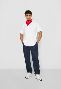 Hvit t-skjorte med svart logo, marineblå cargo-bukser og rød bandana. Modellen har på seg hvite og grå sneakers med svarte detaljer. Enkelt, uformelt antrekk.