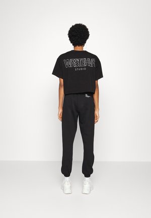 Osoba o krótkich kręconych włosach, ubrana w czarny krótki t-shirt z napisem „WRSTBHVR STUDIO”, czarne spodnie dresowe i białe trampki, stojąca tyłem.