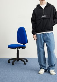 Black windbreaker with a front pocket and "Jordan" logo, paired with light blue wide-leg jeans and white sneakers. Blue office chair beside.