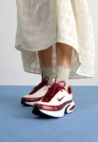 Nike sportbačiai su balta viršutine dalimi, bordo akcentais ir išskirtiniu storu padu. Nešiojami su šviesių spalvų, rengtomis kojinėmis iki kulkšnių ir raštuota sijonu.