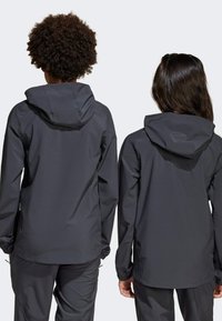 Vestes imperméables noires avec capuches, présentant une texture lisse et mate ainsi que des poignets élastiques, vues de dos sur deux individus.