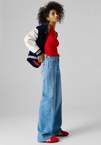 Žena s kudrnatými vlasy, oblečená v červeném tílku, světle modrých džínách širokých nohavic, červených teniskách a nad jedním ramenem má námořnicky modrou bundu typu varsity s bílými prvky.