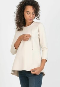 Beige, long-sleeve top with a curved hem and a front slit detail. Made from a smooth fabric, it features subtle stitching and is worn over dark jeans.