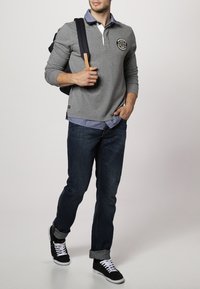 Graues Poloshirt mit langen Ärmeln und weißem Kragen sowie Logo, getragen über einem hellblauen Hemd, kombiniert mit dunklen Jeans und schwarzen Sneakern.