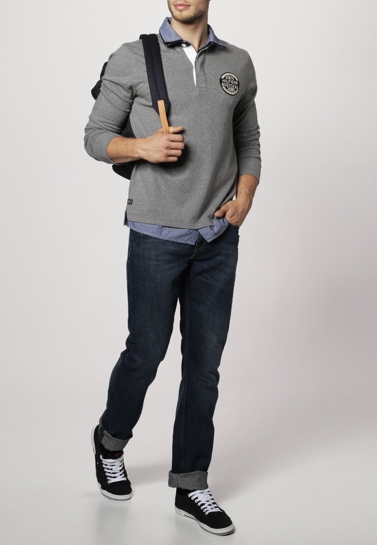 Graues Poloshirt mit langen Ärmeln und weißem Kragen sowie Logo, getragen über einem hellblauen Hemd, kombiniert mit dunklen Jeans und schwarzen Sneakern.