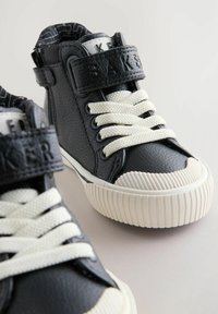 Mustad must kõrged tossud tekstureeritud nahast, valged paelad ja Velcro rihm. Omab ribakujulist valget kummitalda ja logo rihmal.