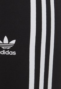 Black athletic pants with three vertical white stripes and a white logo. The material appears soft and stretchy with a smooth texture.