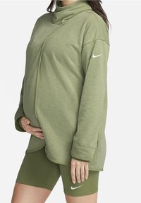 Femme enceinte portant un haut à manches longues style cache-cœur vert clair et un short Nike vert olive, main reposant sur le ventre.