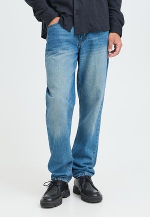 Man draagt lichtblauwe rechte jeans, zwarte veterboots en een donker overhemd met lange mouwen, met één hand in de zak.