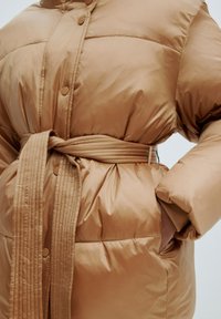 Beige Pufferjacke mit tailliertem Schnitt, ausgestattet mit einer glatten Textur, Druckknopfverschluss und seitlichen Taschen, konzipiert für Wärme und Komfort.