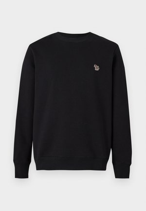 Junger Mann steht, trägt einen schwarzen Sweatshirt und passende schwarze Jogginghosen mit weißen Sneakers vor einem schlichten hellen Hintergrund.