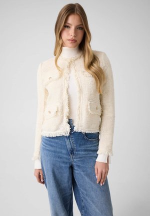 Jeune femme portant une veste texturée crème avec des bords effilochés, par-dessus un col roulé blanc et un jean bleu taille haute, sur un fond uni.