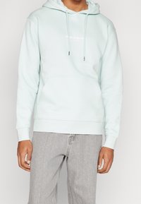 Ljusblå hoodie med framficka, dragsko i huvan och vitt logotryck på bröstet. Har ribbstickade muddar och midjeband.