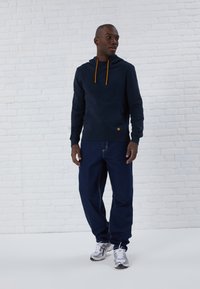 Sudadera navy con cordones naranjas, bolsillo canguro y parche de logo. Combinada con jeans azules y zapatillas deportivas plateadas sobre un fondo claro.