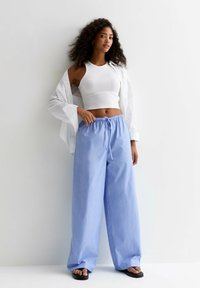 Pantalons larges rayés bleu clair avec une taille élastique, assortis d'un crop top blanc ajusté et d'une chemise blanche ouverte. Sandales plates portées.