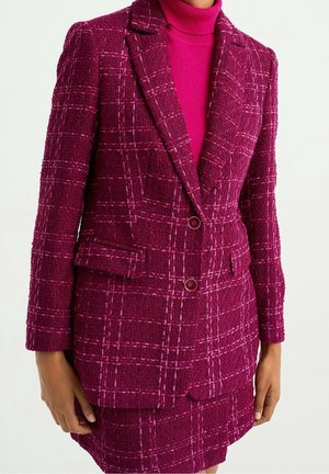 Donna che indossa una giacca a quadri rosa scuro con trama e bottoni, sopra un dolcevita rosa coordinato e una gonna, in piedi con le braccia rilassate lungo i fianchi.