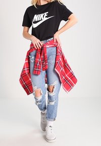 Schwarzes Nike-T-Shirt mit weißem Logo, gealterte hellblaue Jeans, rot kariertes Hemd um die Taille gebunden und weiße Turnschuhe.