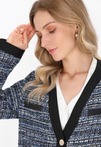 Blazer a quadri blu e nero con colletto nero e due tasche frontali, caratterizzato da un bottone dorato e una camicetta bianca liscia sotto.