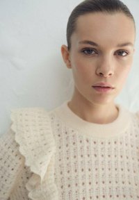 Suéter de punto color crema con un patrón texturizado, cuello redondo acanalado y un detalle de volantes en el hombro izquierdo. Acabado suave.