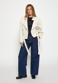 Chaqueta corta de color beige claro con solapas anchas y mangas largas, llevada sobre una blusa negra y combinada con unos jeans oscuros de pierna ancha y talle alto.