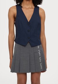 Marinblå väst med djup urringning och fyra svarta knappar, i kombination med en grå plisserad kjol med vertikala ränder och vit text.