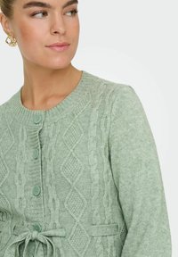 Svetlo zelena jopica z zasnovo iz teksturirane diamante in kabelskim pletenjem. Ima rebrast ovratnik in sprednje gumbke ter pas s kravato.