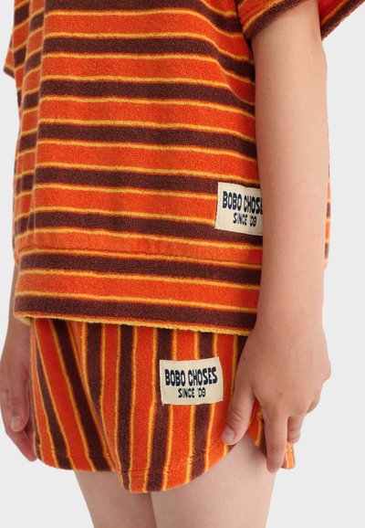 Enfant portant une chemise et un short en éponge rayés orange et marron, tous deux étiquetés "BOBO CHOSES DEPUIS 09".