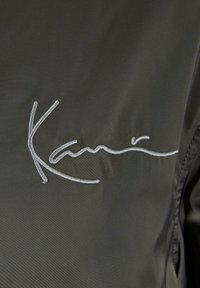 Schwarzer Stoff mit einer glatten Textur, der mit weißen gestickten Signaturdetails verziert ist. Das Design ist minimalistisch und konzentriert sich auf die Umrisse des Logos.