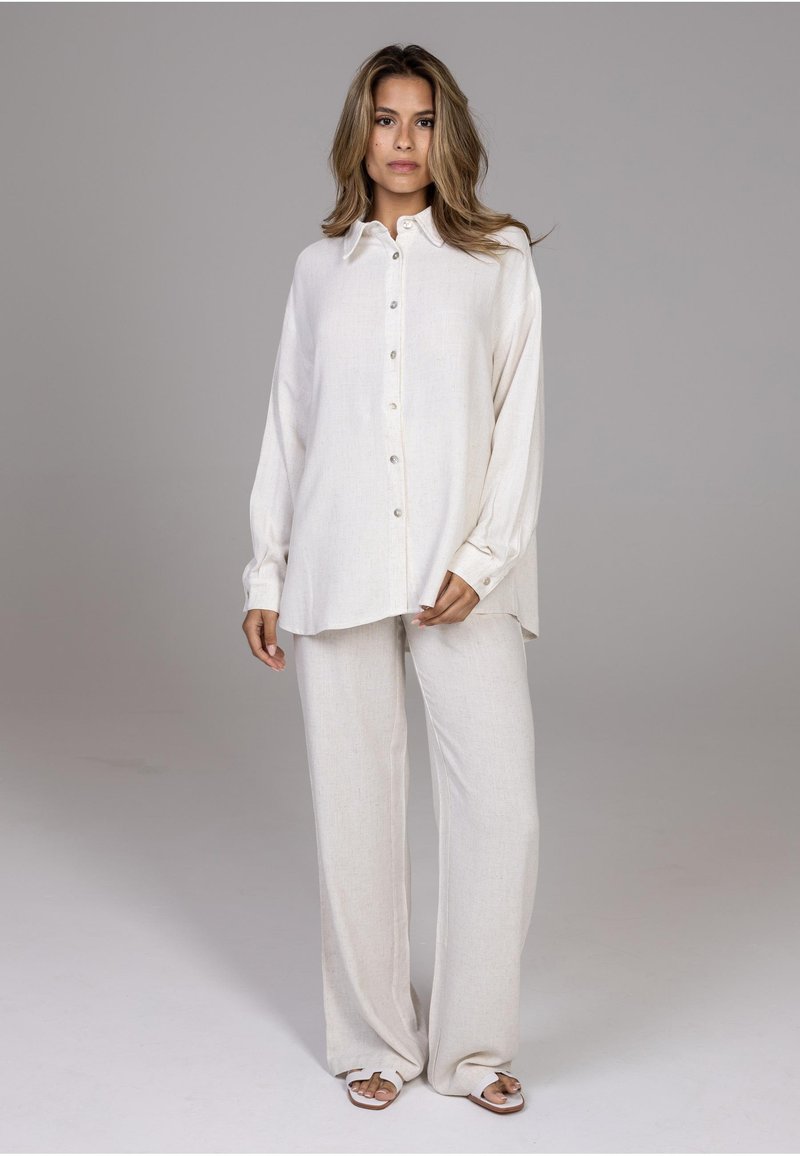 Femme debout portant une chemise blanche ample boutonnée, un pantalon à jambes larges et des sandales ouvertes sur un fond gris uni.