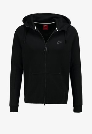 Ein Mann geht, trägt eine schwarze Nike-Zip-up-Hoodie, ein weißes T-Shirt, graue Jeans und weiße Turnschuhe vor einem einfarbigen Hintergrund.