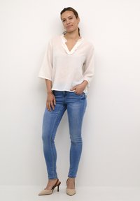 Bluză ușoară, vaporoasă, cu guler volan, asortată cu blugi aderenti albaștri și pantofi cu toc înalt bej. Textură netedă și stil casual.