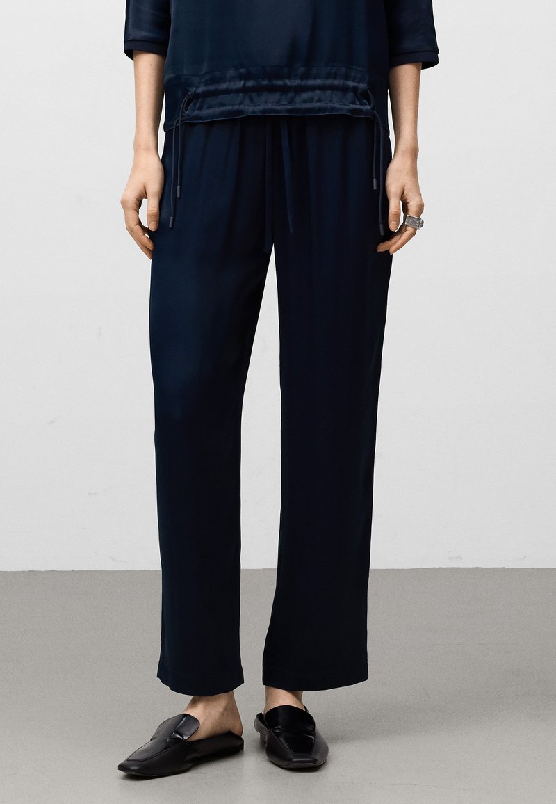 Person som bär lösa marinblå byxor och svarta slip-on skor, står på ett helt grått golv mot en vit vägg.