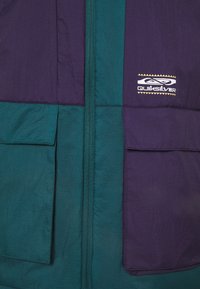 Veste violette et bleu sarcelle à l'avant avec une fermeture éclair centrale, deux grandes poches à rabat, et le logo blanc Quiksilver sur le côté supérieur droit.