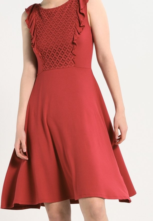 Robe rouge sans manches avec motif au crochet et volants sur la poitrine, taille cintrée et jupe évasée tombant au-dessus des genoux.