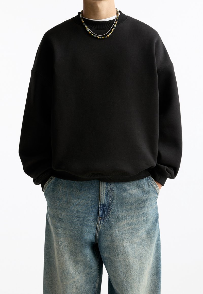 Personne portant un sweat-shirt noir ample, un jean bleu avec les mains dans les poches, et des colliers de perles superposés sur un fond blanc uni.