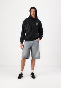 Black hooded sweatshirt with logo, blue denim shorts, and black sneakers. Accented with a small black crossbody bag. Plain white background.