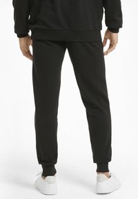 Pantalones de chándal negros hechos de una tela suave con puños y cintura acanalados. Combinados con zapatillas blancas que tienen un logo en el talón.