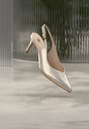 Chaussure à talon haut slingback dorée métallique avec bout pointu et sangle à boucle, flottant au-dessus d'un sol en béton poli.