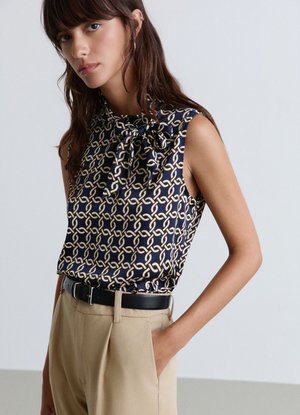 Donna che indossa una camicetta senza maniche blu navy con motivo a catena beige e fiocco, abbinata a pantaloni beige a vita alta e cintura nera.