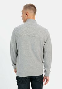 Sweatshirt gris avec une texture matelassée sur le haut du dos, un col montant et des poignets côtelés. Porté avec un jean sombre, affiché de dos.