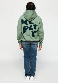 Sweat à capuche vert avec un grand lettrage vert foncé "MULTPLY" dans le dos. Associé à un jean bleu et des baskets blanches. Design et coupe décontractés.